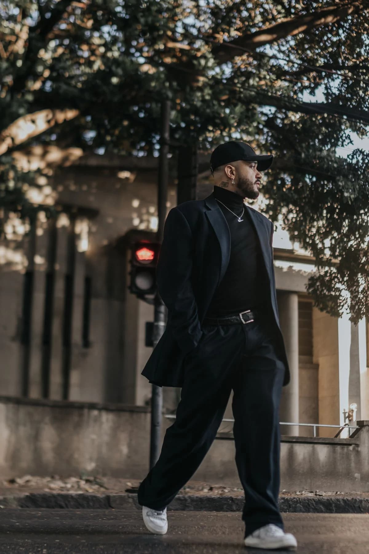 Full-length style portrait: black tailoring and turtleneck, relaxed trousers, cap, pendant and white sneakers