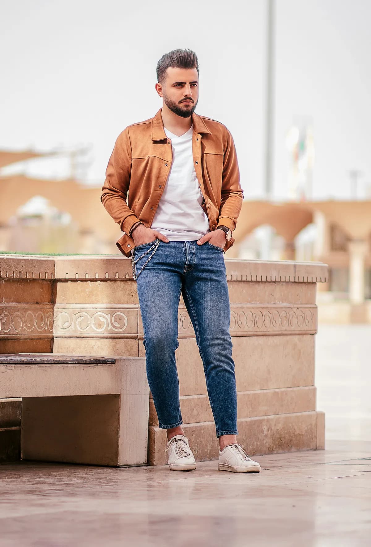 Man in a tan jacket, white T-shirt, blue jeans and white sneakers beside a stone bench